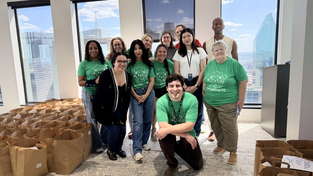 A large group of volunteers in Kind Cooking t-shirts gathered beside rows of assembled food bags.